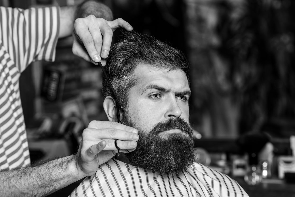 Barber man serving client at barber shop. Vintage barbershop, shaving. Brutal bearded guy viziting hairstylist in barber shop. Making haircut look perfect. Hairdresser, hair salon. Black and white.